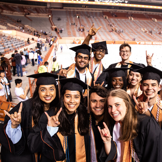 Group of graduates smile and give hook em horns in caps and gowns